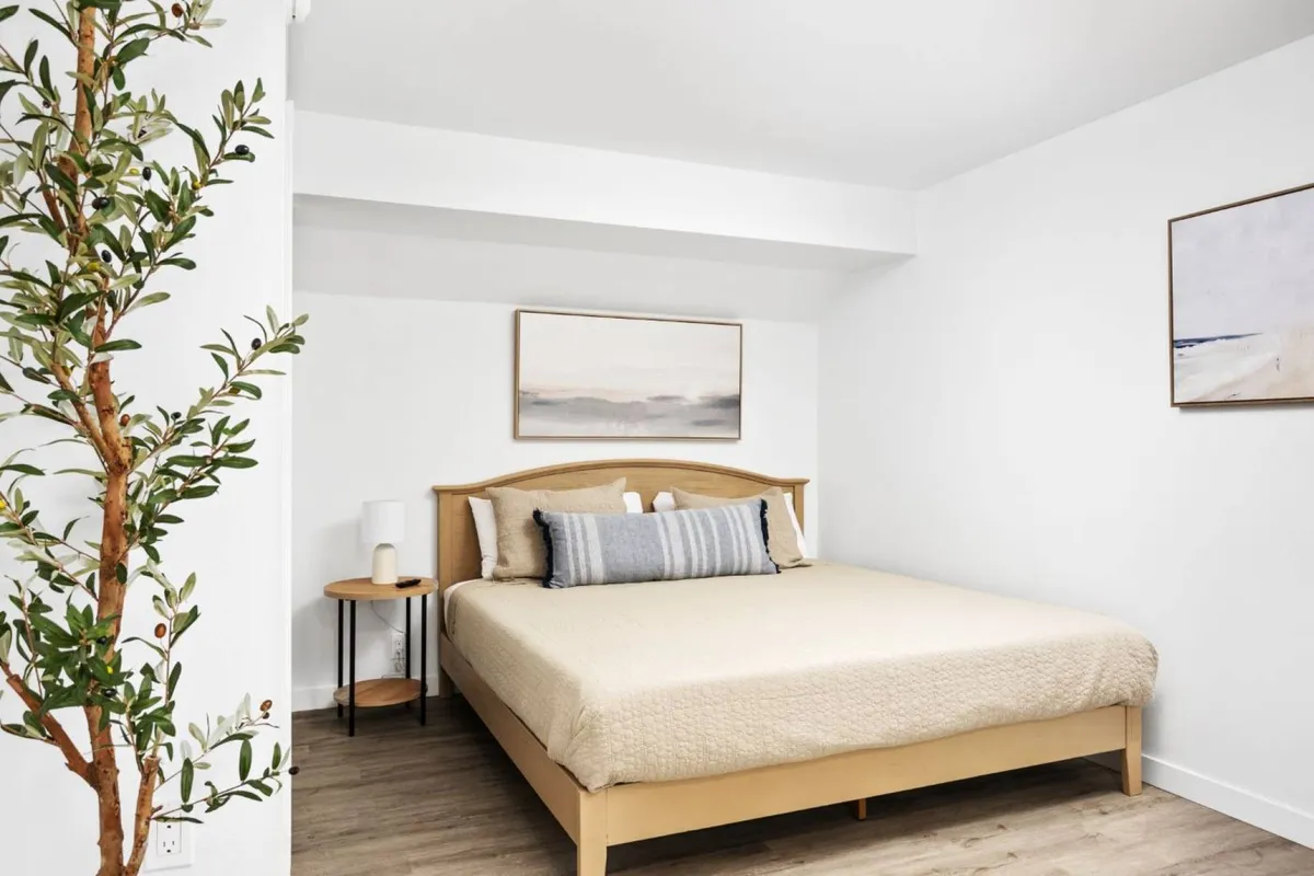 A bright and peaceful corner of the studio with a well-made bed, decorative artworks on the wall, a side table with a leafy plant, and soft natural lighting, offering a sense of private sanctuary.