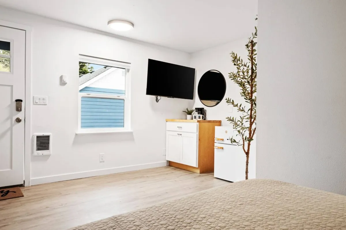 This interior image features a comfortable, modern studio space with a large window providing a view of the outdoors. The scene includes a TV mounted on the wall, a round mirror, a small potted plant, and a wooden dresser, all arranged to create a serene, inviting atmosphere. Soft natural light fills the room, suggesting the proximity to the coast and evoking the relaxed mornings described in the section.