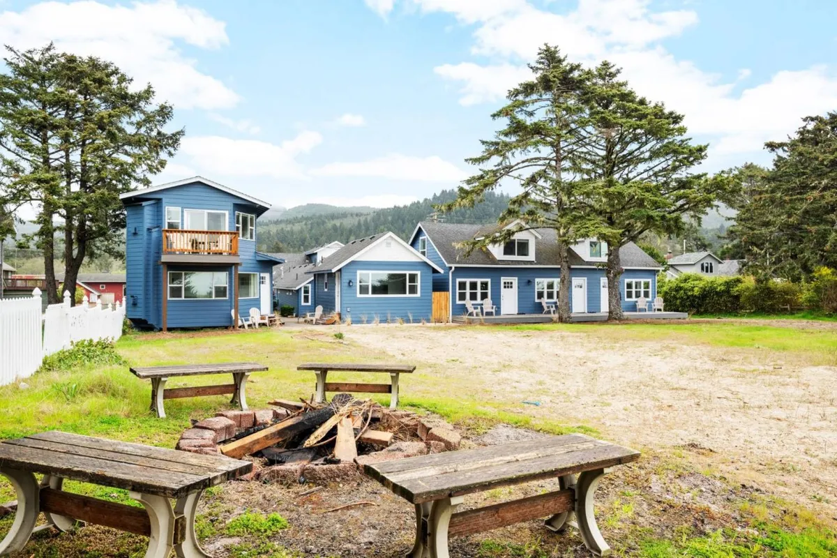 A welcoming outdoor communal fire pit area surrounded by green lawn and framed by the blue Ocean Rogue Inn houses. Several chairs are set around the fire pit, inviting guests to gather, share stories, and enjoy the warmth. Mature trees in the background add a touch of natural serenity to the communal space.