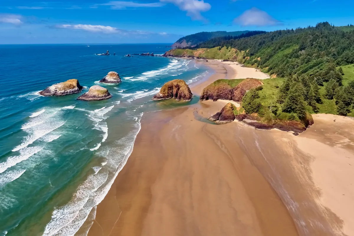 A tranquil scene capturing the sandy beach bordered by lush green hills and striking rock formations. Waves gently lap at the shore, and the light hints at an impending sunset. The natural scenery suggests opportunities for exploration, beachcombing, and enjoying the outdoors.