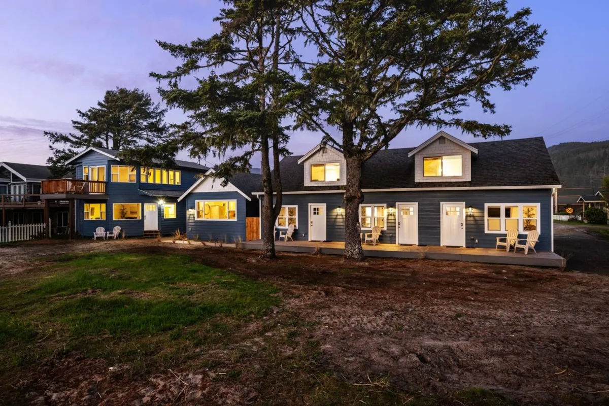 The exterior of a charming two-story condo is nestled among tall trees, with glowing windows that suggest warmth and comfort inside. A welcoming outdoor seating area sits beneath the trees, perfect for relaxing or gathering with friends, and the overall scene exudes an inviting and cozy atmosphere, ideal for enjoying sunsets or meals outdoors.